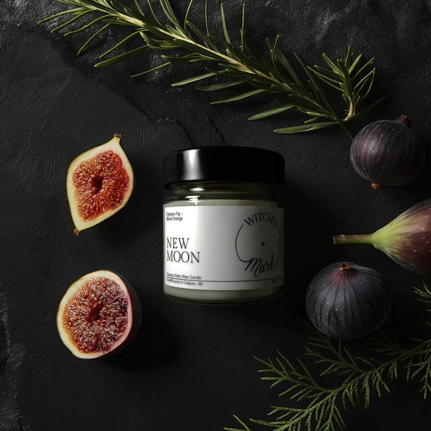 A top-down flat lay highlighting the scent profile of the New Moon candle, featuring the jar surrounded by fresh halved figs and sprigs of green cypress on a dark slate background.