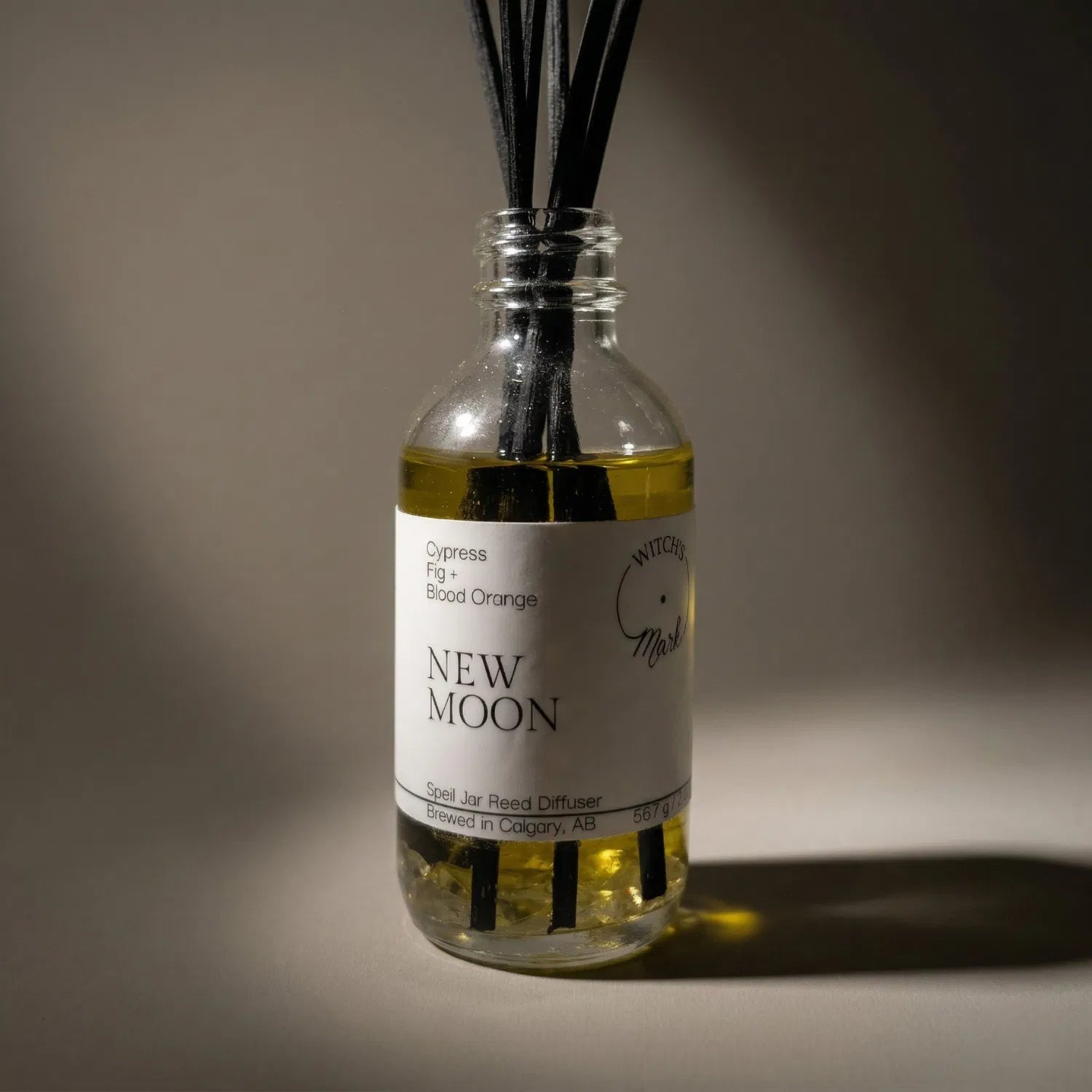 A dramatic spotlight close-up of the New Moon spell jar reed diffuser, highlighting the front label which lists its magical, energetic scent profile of grounding cypress, fig, and bright blood orange.