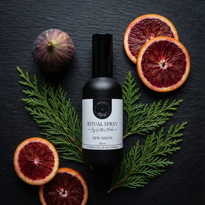 A top-down flat lay highlighting the invigorating ingredients of the New Moon ritual mist by Witch's Mark. The black apothecary bottle sits surrounded by fresh green cypress sprigs, a ripe whole fig, and slices of bright blood orange against a dark slate background.