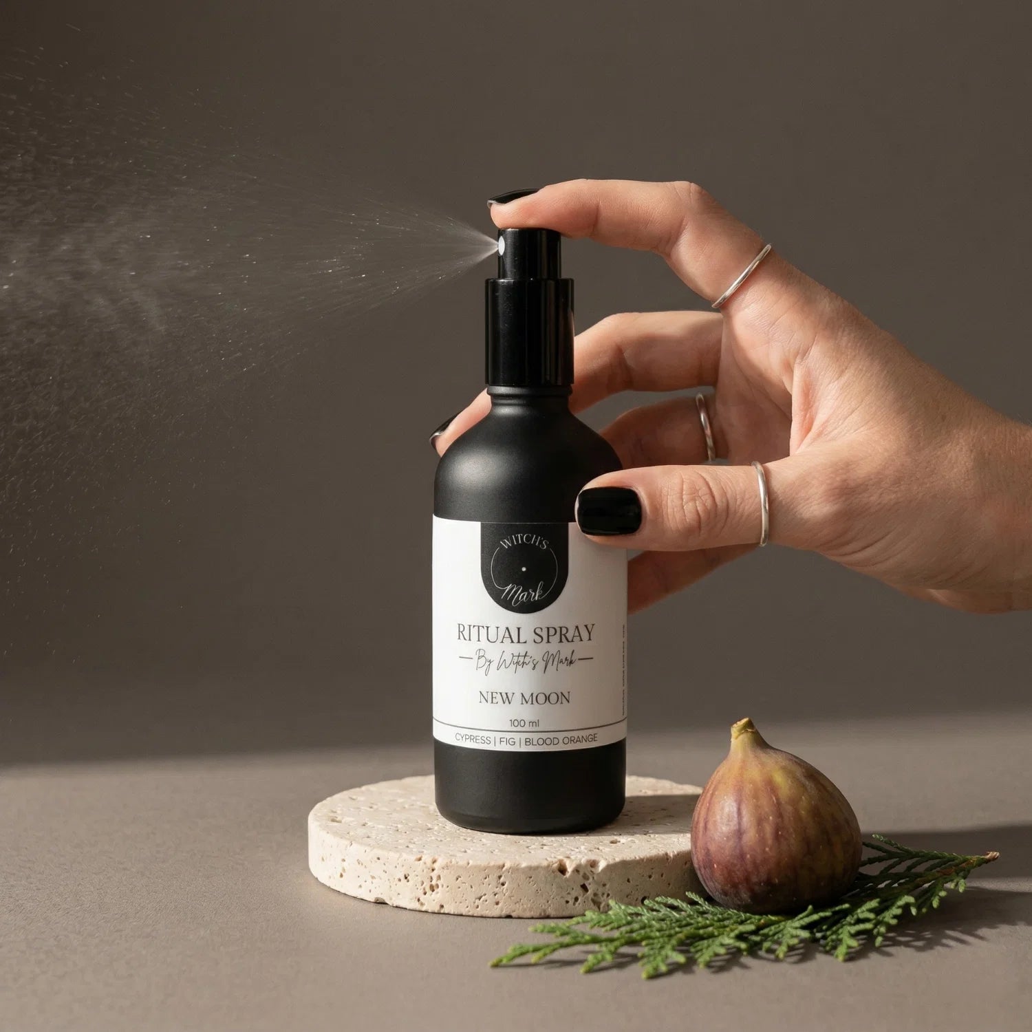 A hand with black nail polish spritzing the Witch's Mark New Moon ritual spray over a fresh whole fig and a sprig of green cypress. This awakening aura mist is infused with clear quartz charged moon water to clear the slate and plant energetic seeds for profound new beginnings.