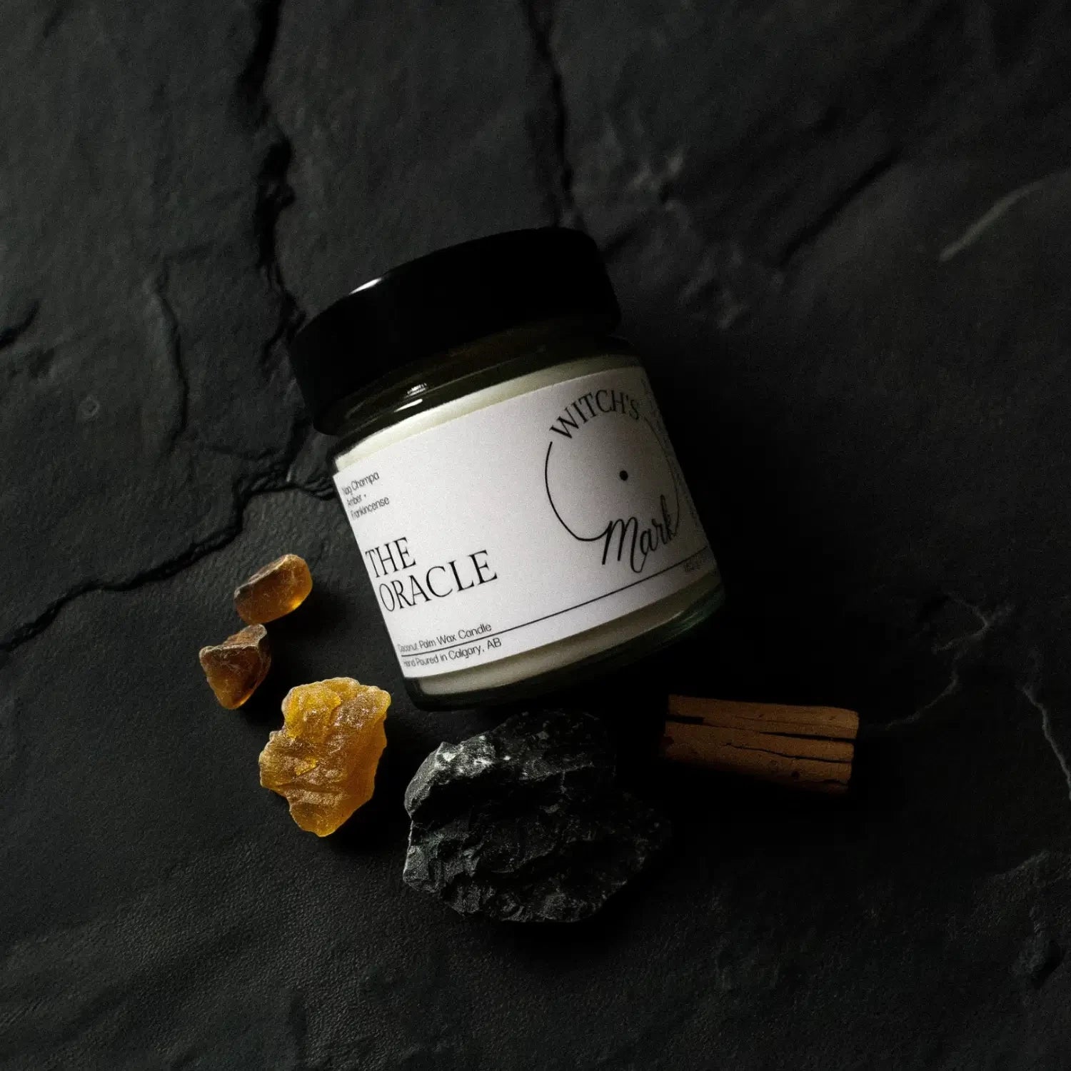 A top-down flat lay highlighting the ingredients of The Oracle candle, featuring the jar surrounded by raw chunks of warm amber and natural frankincense tears on a dark slate background.