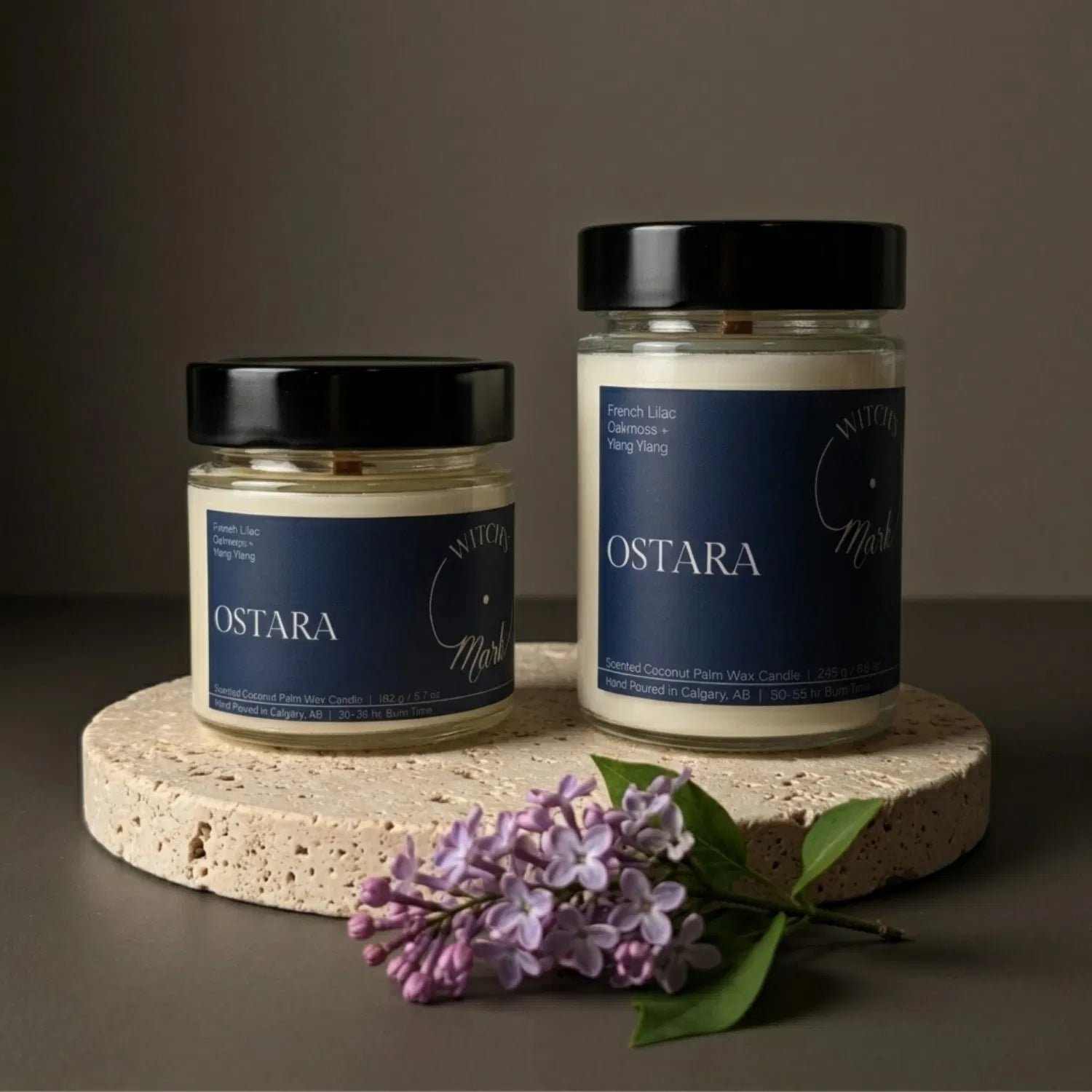 Two sizes of the seasonal Witch's Mark Ostara coconut wax candle, 5.7 oz and 8.6 oz, displayed on a round stone base and garnished with fresh purple French lilac blooms.