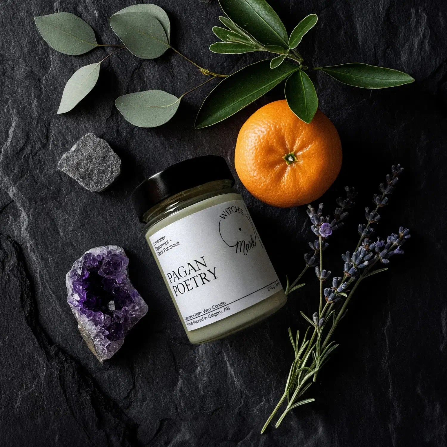 A top-down flat lay highlighting the ingredients of the Pagan Poetry candle, featuring the jar surrounded by a raw amethyst crystal, fresh lavender, spearmint leaves, and citrus on a dark slate background.