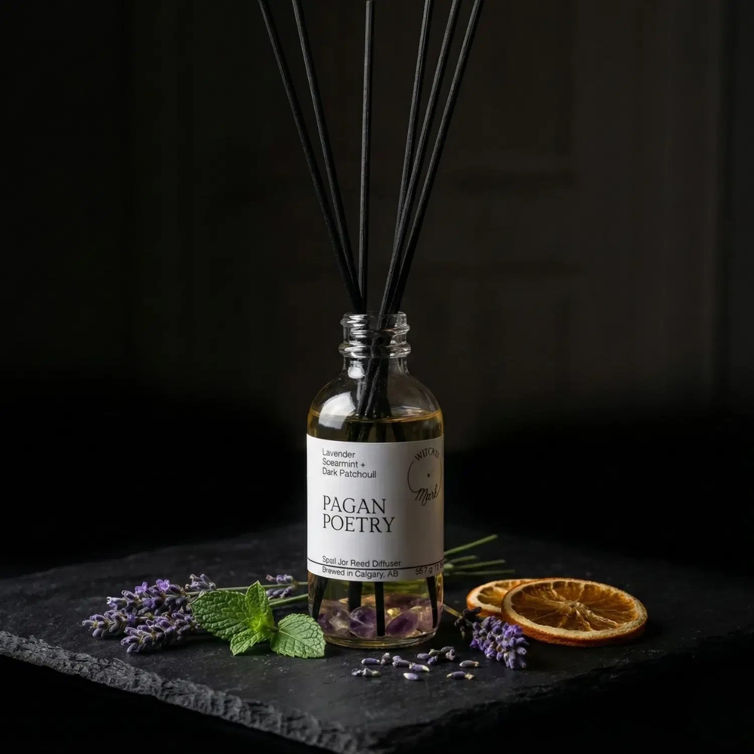 The Pagan Poetry spell jar reed diffuser by Witch's Mark showcased against a dark slate background, surrounded by its core botanical ingredients: fresh purple lavender, crisp green spearmint leaves, and earthy botanical elements.
