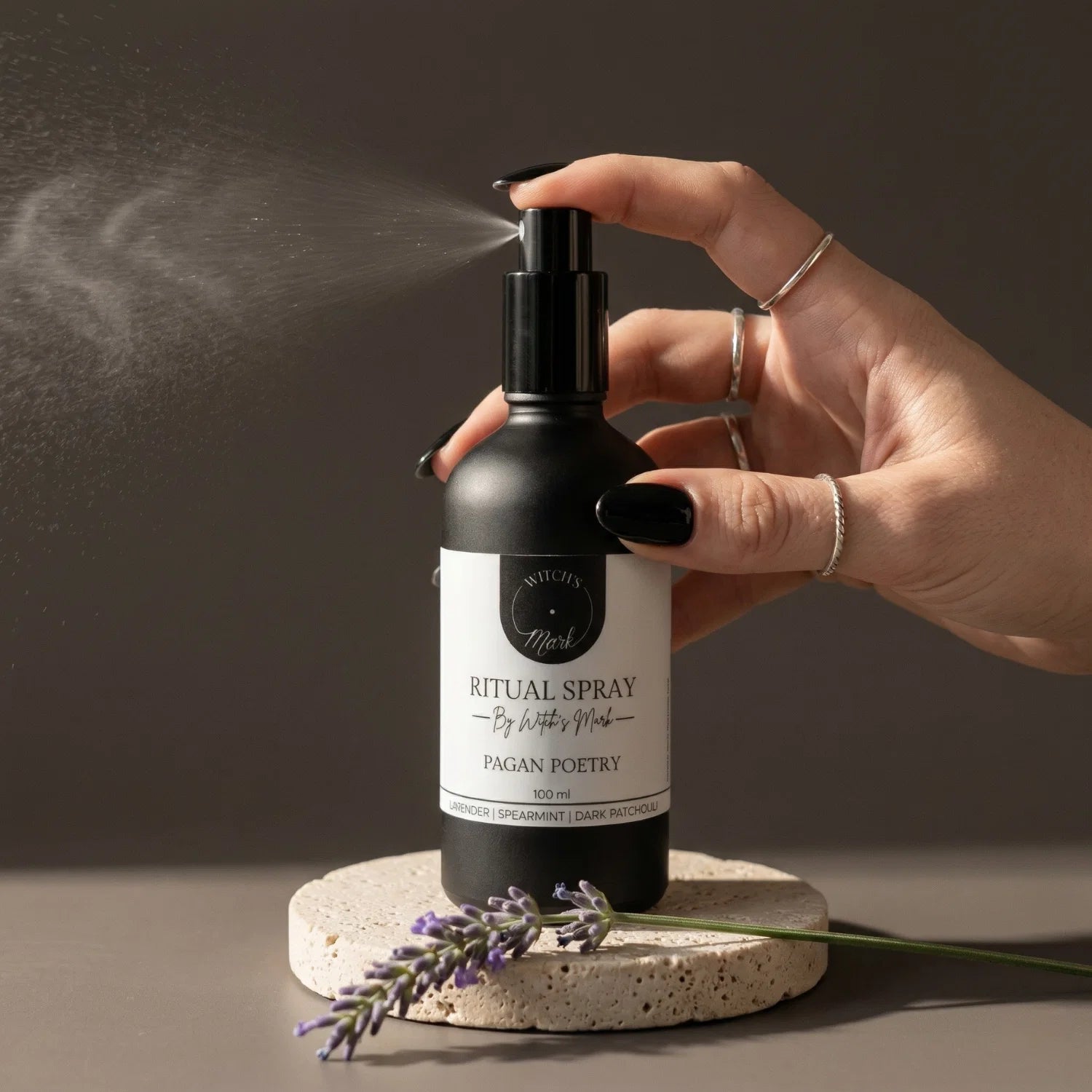 A hand with black nail polish spritzing the Pagan Poetry ritual spray by Witch's Mark over a sprig of fresh lavender, designed for instant atmospheric shifts and energy restoration.