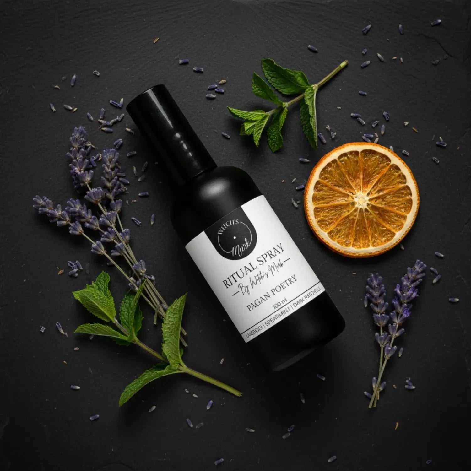 A top-down flat lay highlighting the restorative ingredients of the Pagan Poetry ritual mist by Witch's Mark, featuring the black apothecary bottle surrounded by fresh calming lavender, crisp spearmint leaves, and a slice of dried citrus against a dark slate background.