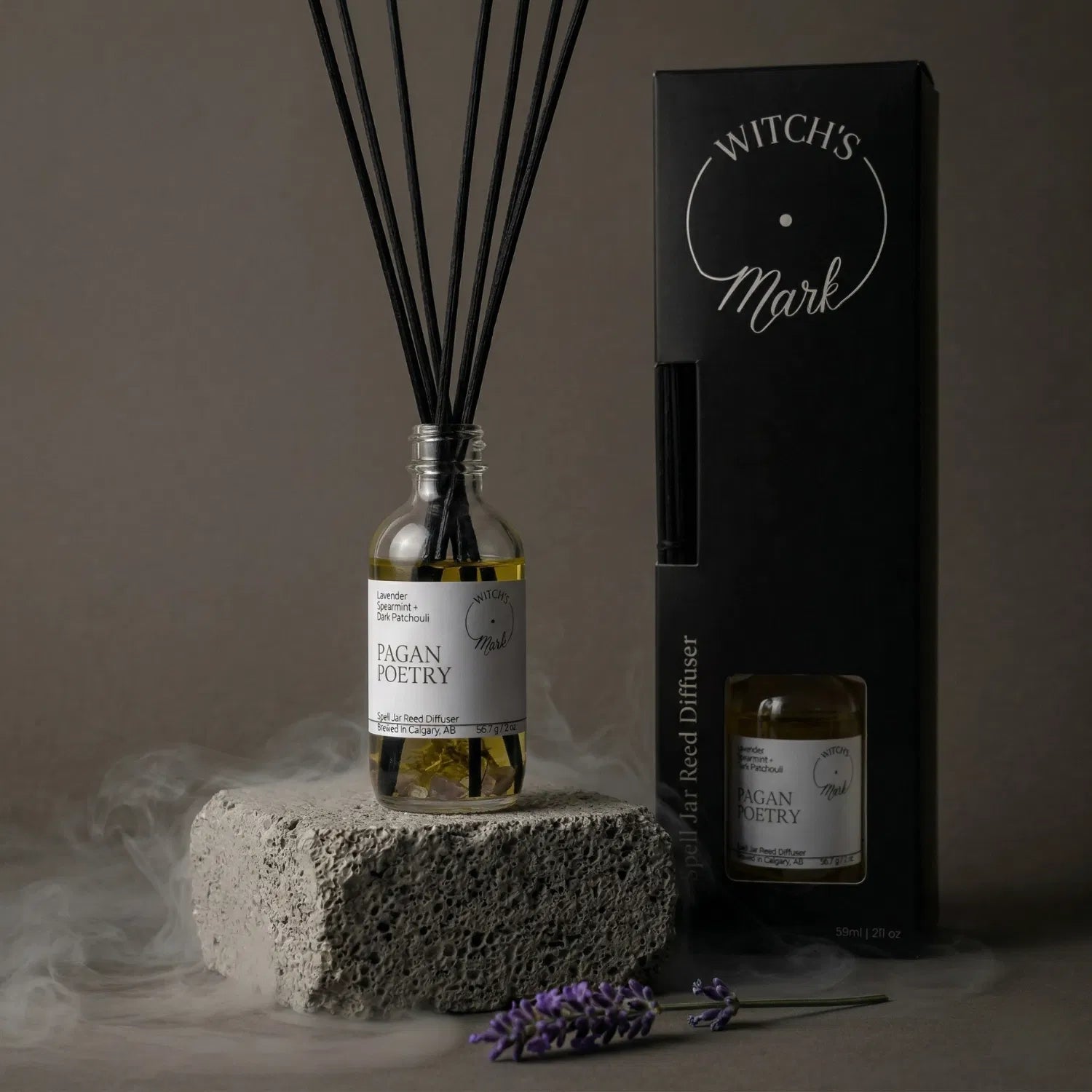 A Pagan Poetry spell jar reed diffuser by Witch's Mark showcased with its dark packaging and fresh lavender, offering continuous calming aromatherapy in a misty setting.