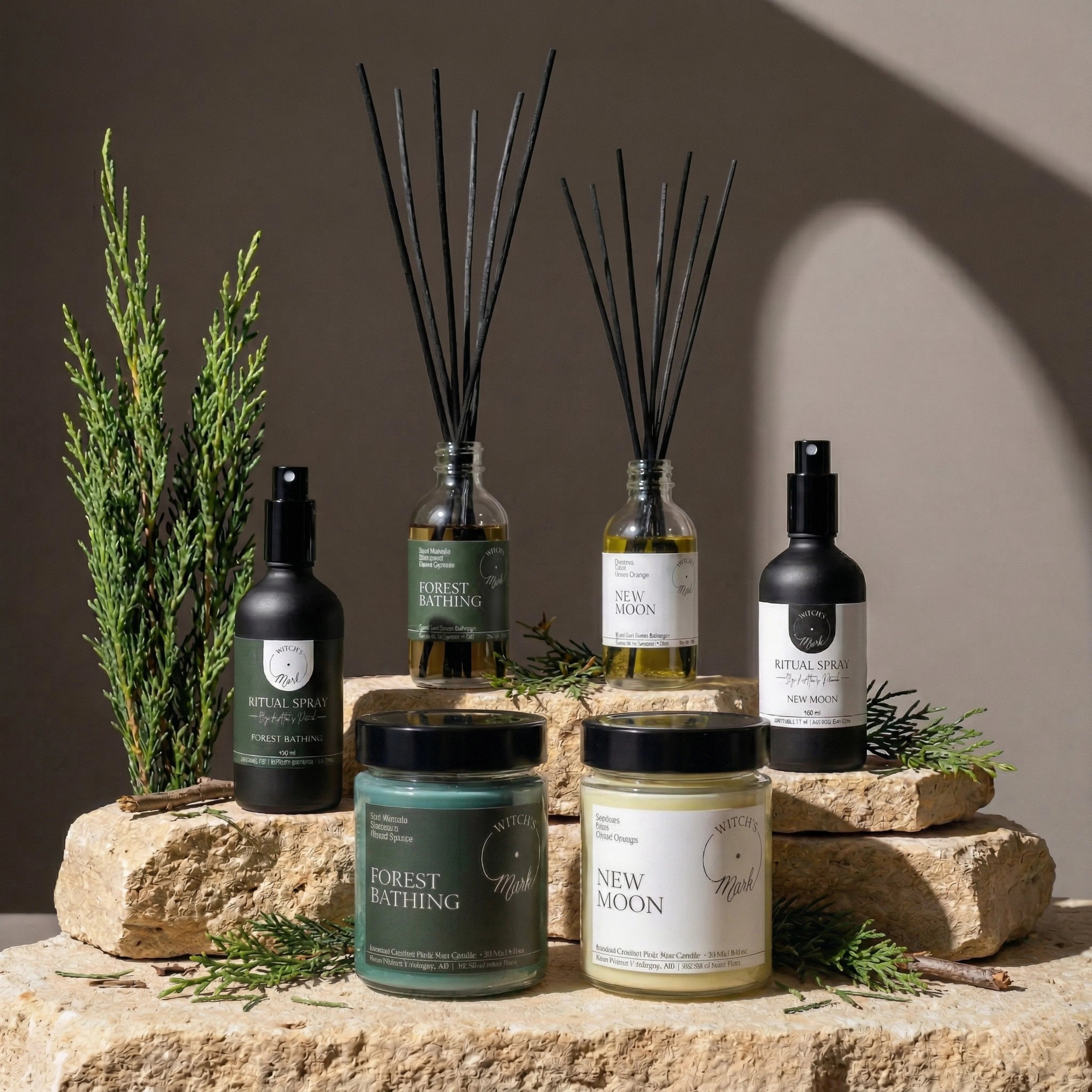 The complete Rooted Intentions Ritual Bundle by Witch's Mark, featuring Forest Bathing and New Moon crystal candles, spell jar reed diffusers, and ritual sprays beautifully arranged on textured stones with fresh woodland greenery.