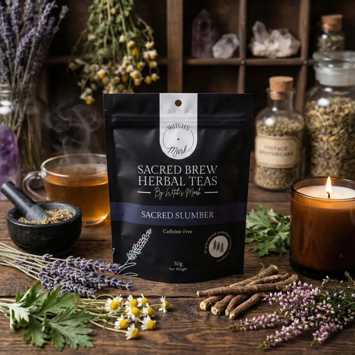 The Sacred Brew Herbal Teas Sacred Slumber pouch by Witch's Mark displayed on a wooden apothecary table. This caffeine-free bedtime tea is surrounded by its raw restorative ingredients including fresh lavender, chamomile, heather flower, mugwort, and valerian root, alongside a lit amber candle and a freshly brewed cup.