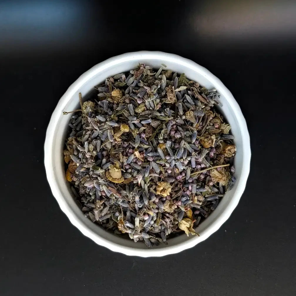 A top-down, close-up view of the Sacred Slumber loose leaf herbal tea blend by Witch's Mark in a small white bowl. The image clearly showcases the high-quality mix of dried lavender buds, chamomile flowers, and restorative nighttime herbs against a dark background.
