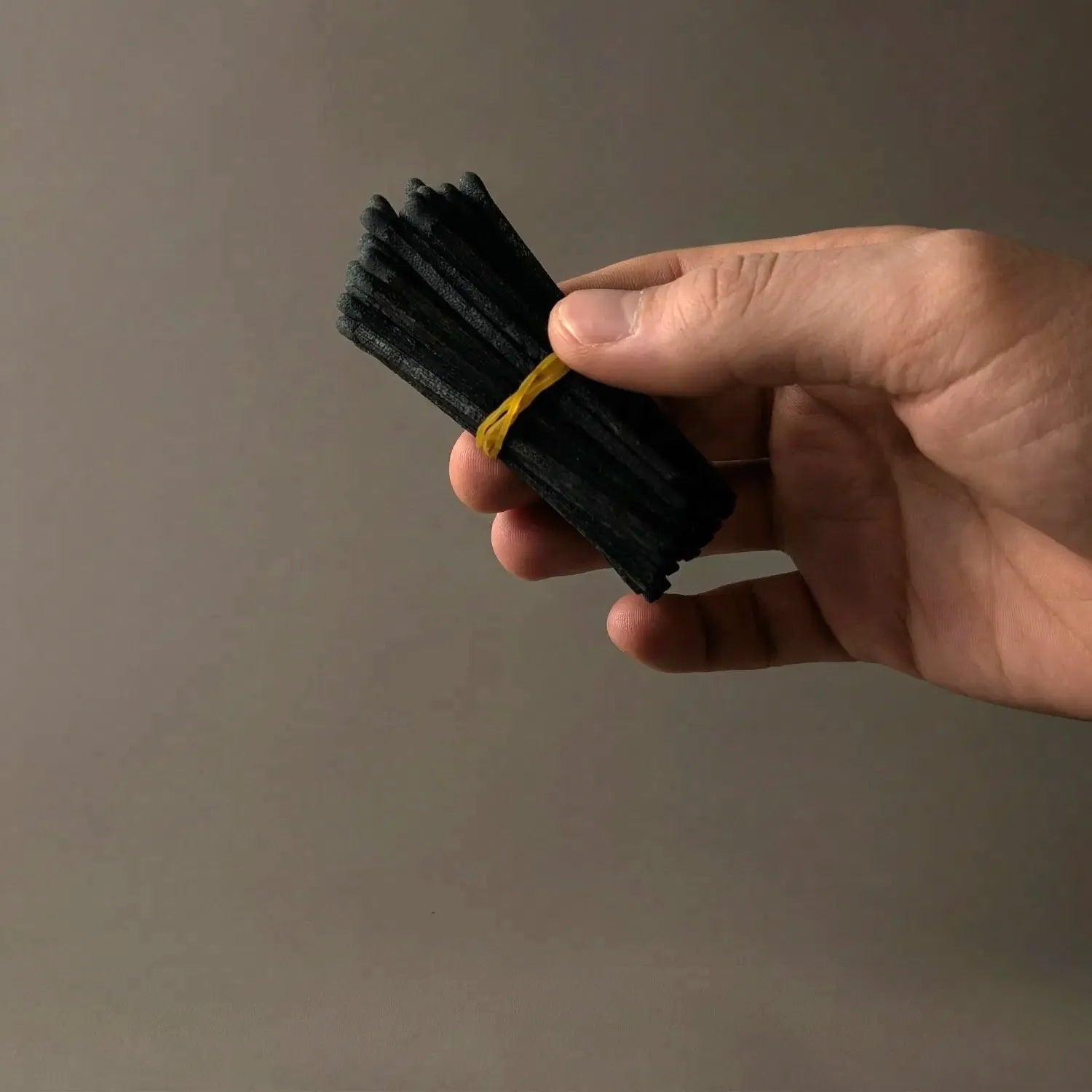 A hand holding a bundled refill of 50 sleek, 3-inch black-on-black matches bound by a yellow band. These dark, aesthetic matches are perfect for restocking your cauldron match holder or filling your own apothecary matchbox.