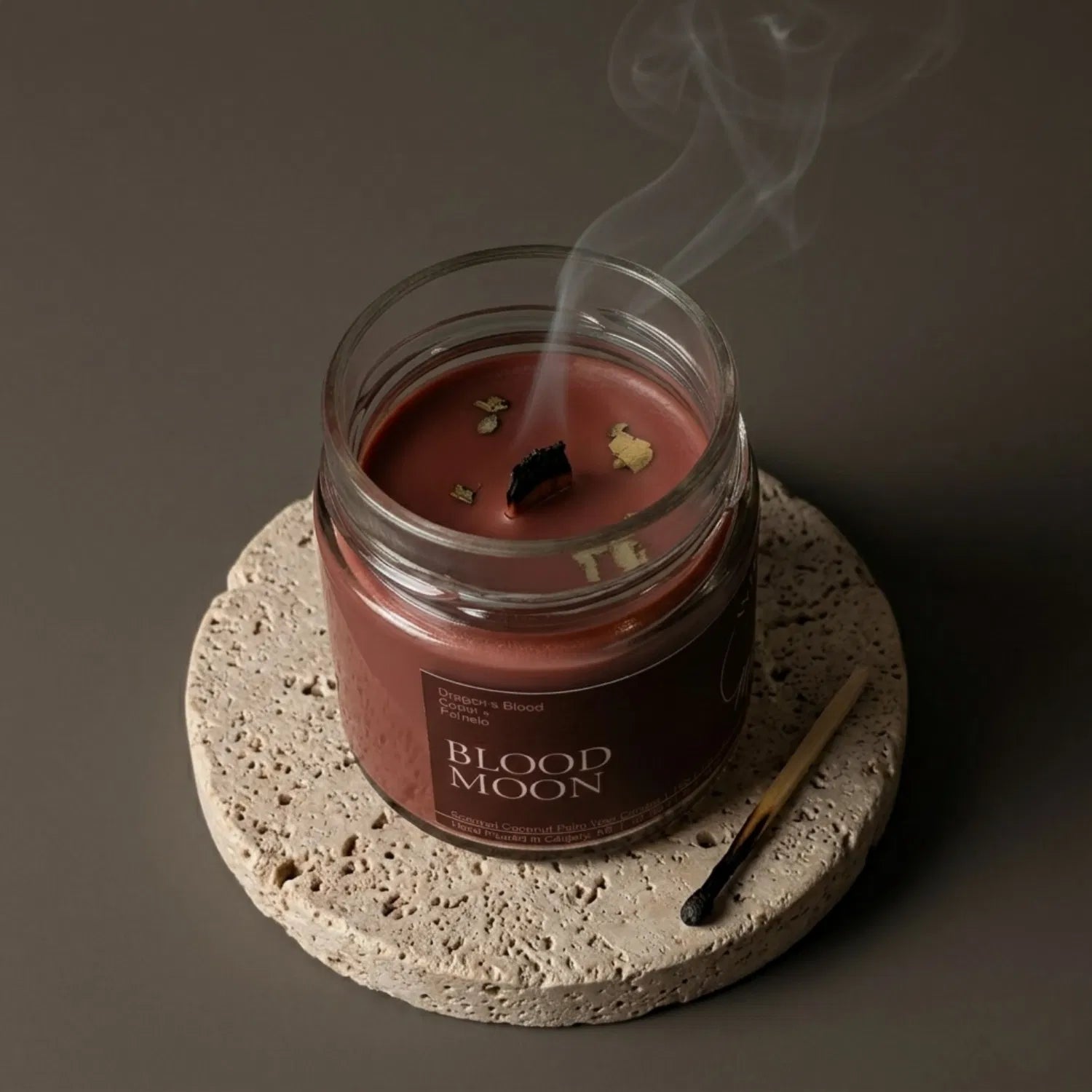 A freshly extinguished Blood Moon candle resting on a stone coaster, emitting a delicate trail of calming smoke from its wooden wick after a healing ritual. The red wax is lightly adorned with energizing yerba mate, with a burnt match resting nearby.