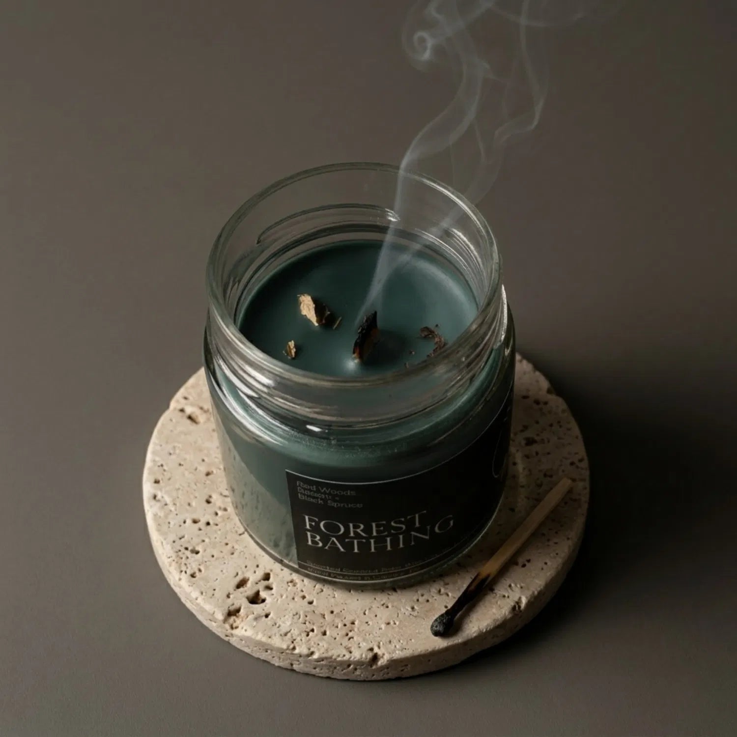 A freshly extinguished Forest Bathing candle resting on a stone coaster, emitting a delicate trail of smoke from its wooden wick after a grounding ritual. The green wax is lightly adorned with lemon balm, with a burnt match resting nearby.