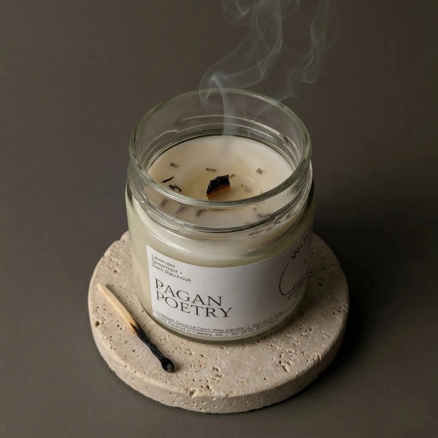 A freshly extinguished Pagan Poetry candle resting on a stone coaster, emitting a trail of calming smoke from its wooden wick, with a burnt match resting nearby.