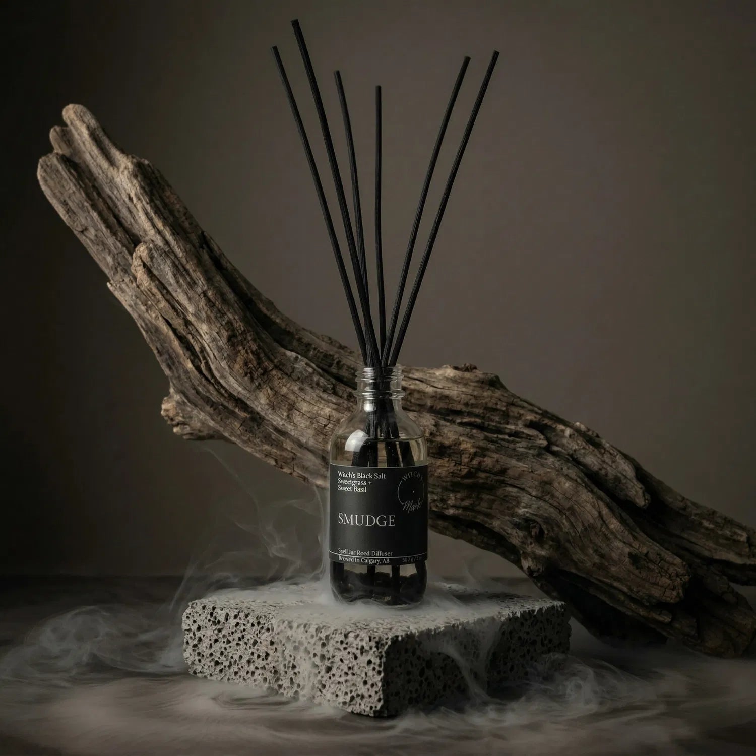 The Smudge apothecary spell jar reed diffuser by Witch's Mark sitting on a stone base, enveloped in atmospheric mist with natural driftwood in the background, designed to act as a continuous smokeless smudge to clear stagnant energy.