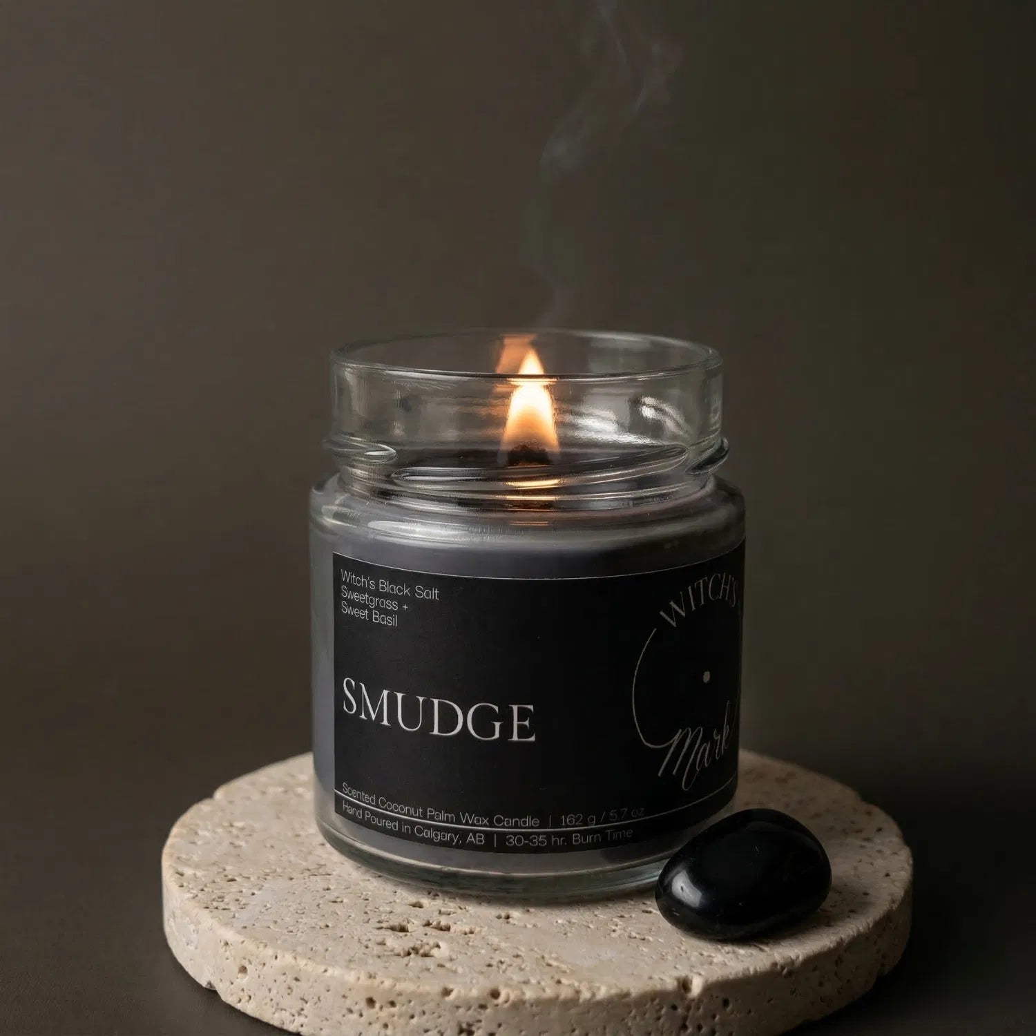 A lit Smudge crystal candle by Witch's Mark sitting on a textured stone coaster next to a black obsidian crystal, with cleansing smoke gently rising against a dark, moody background.