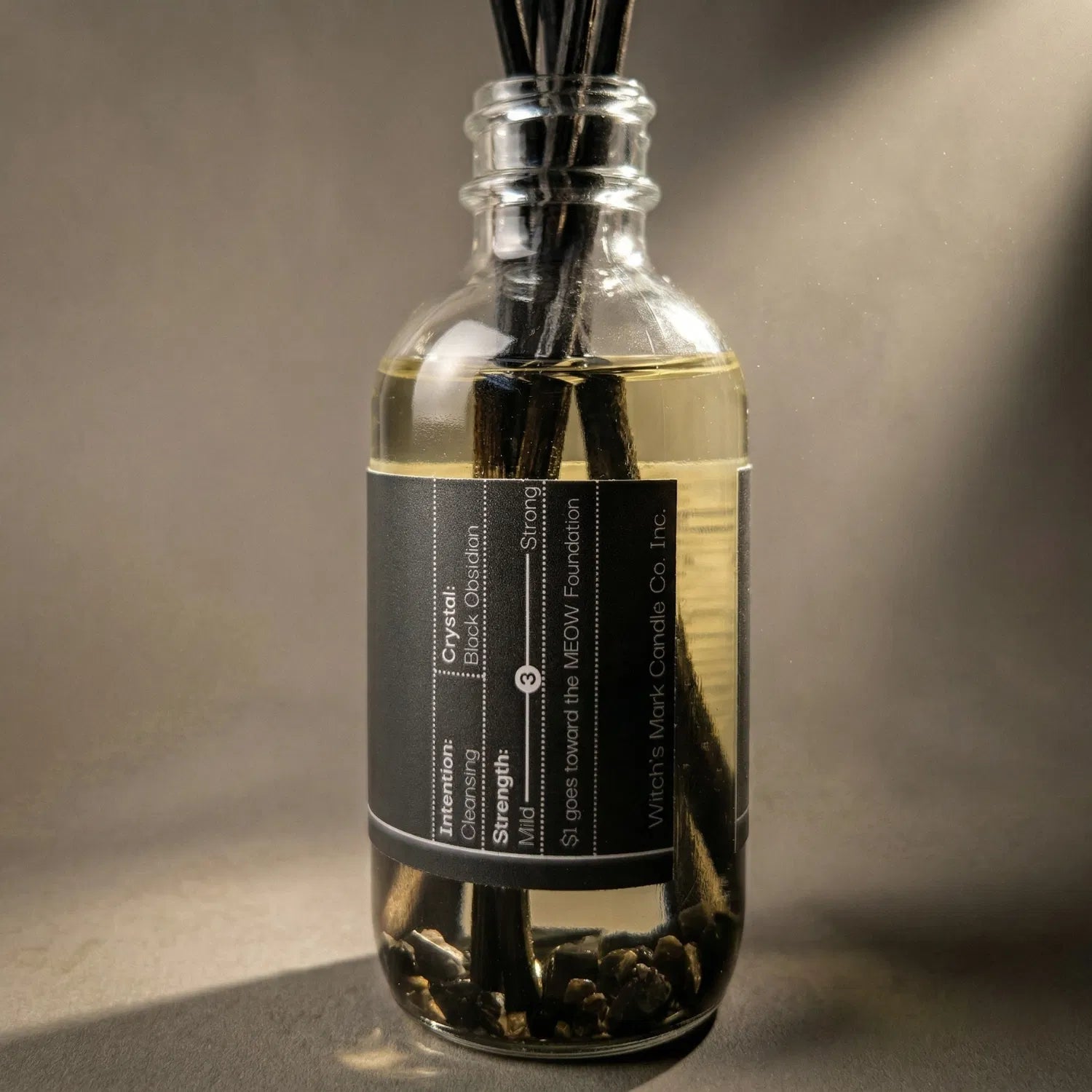 A warmly lit close-up of the Smudge spell jar reed diffuser, revealing the potent black obsidian crystals submerged at the bottom of the clear botanical oil, crafted to absorb profound negativity and provide supreme psychic protection.