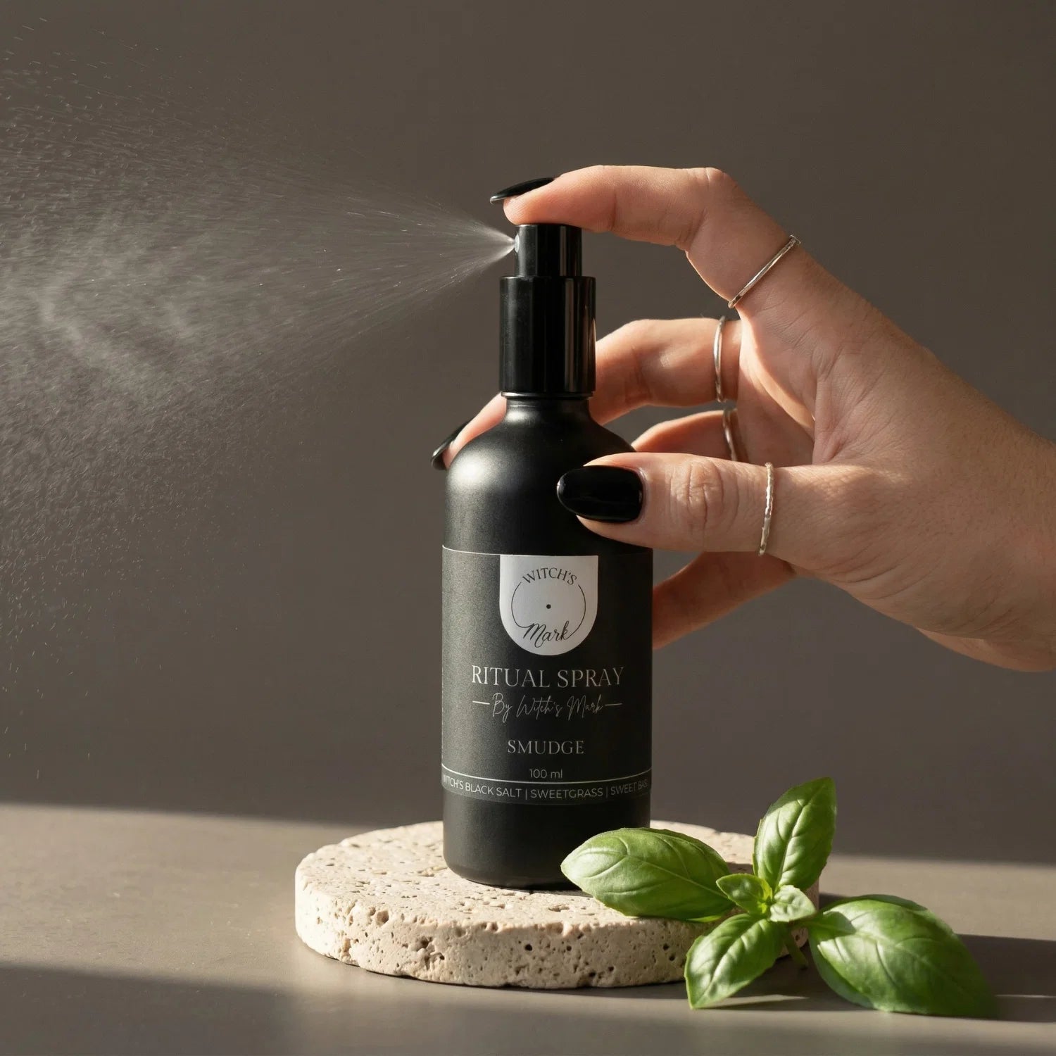 A hand with black nail polish spritzing the Witch's Mark Smudge ritual spray over fresh sweet basil. This smokeless smudge mist is infused with black obsidian charged moon water to banish negative energy and cleanse the aura.