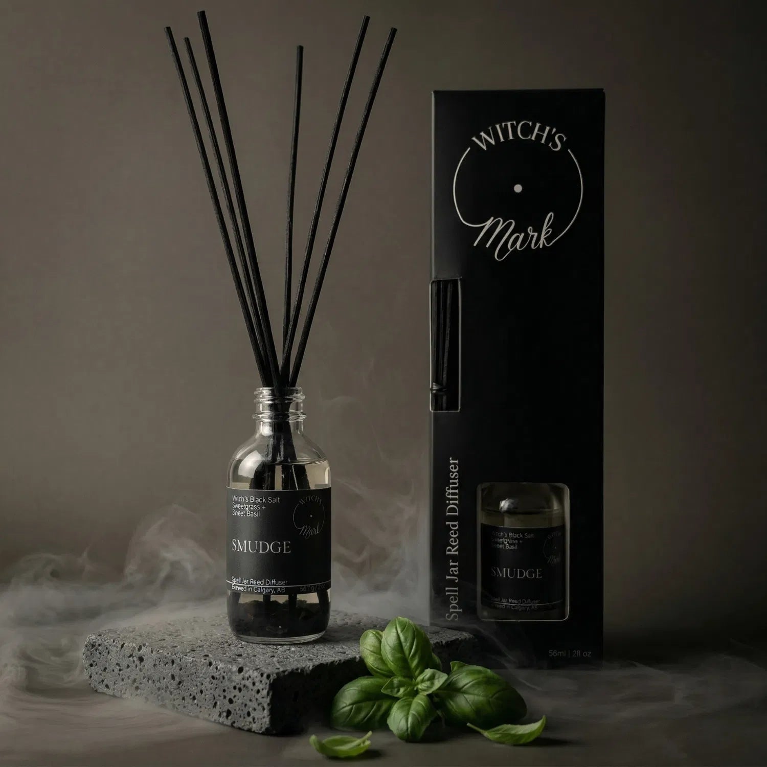 The Smudge spell jar reed diffuser by Witch's Mark displayed next to its sleek black packaging box. The apothecary glass vessel with black reeds rests on a textured stone block surrounded by mystical mist and fresh sweet basil leaves, serving as a perfect smokeless space clearing tool.