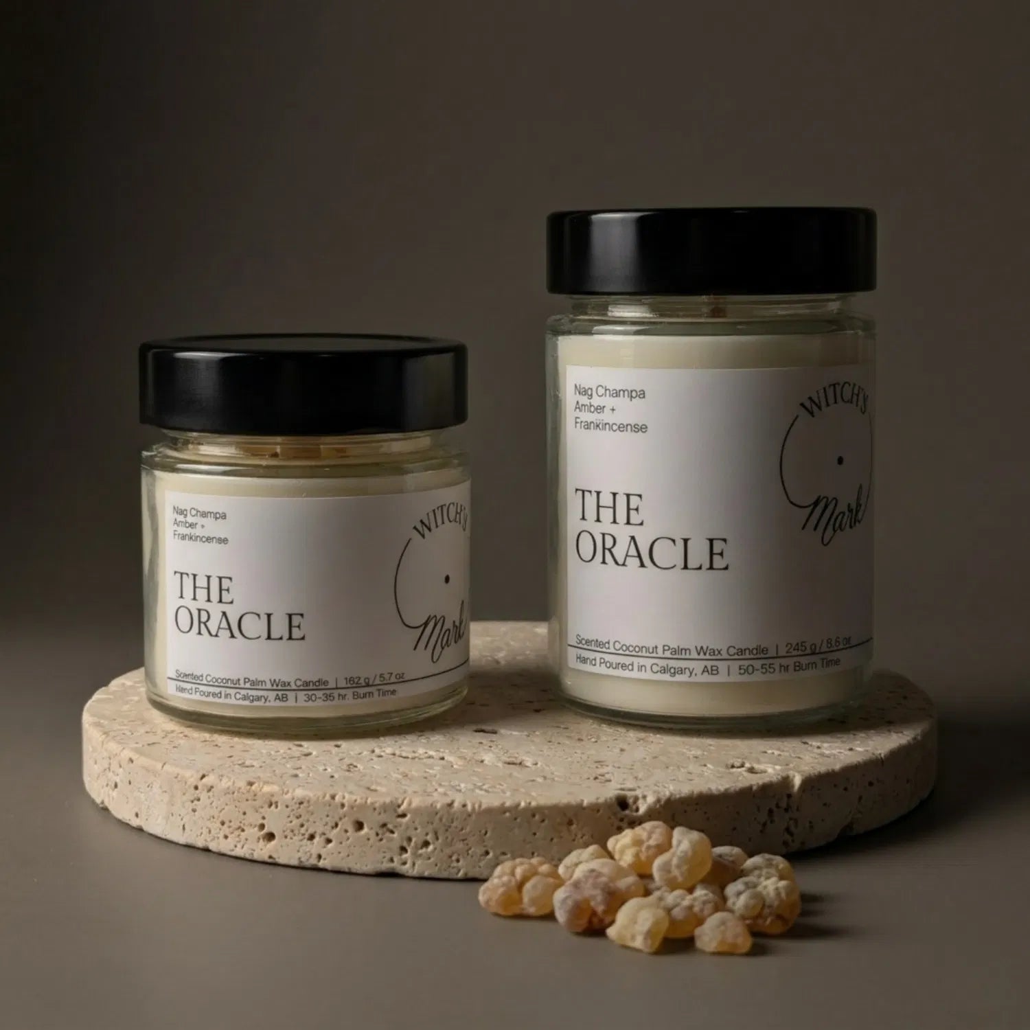 Two sizes of the Witch's Mark Oracle coconut wax candle, 5.7 oz and 8.6 oz, displayed on a round stone base and garnished with natural frankincense tears.