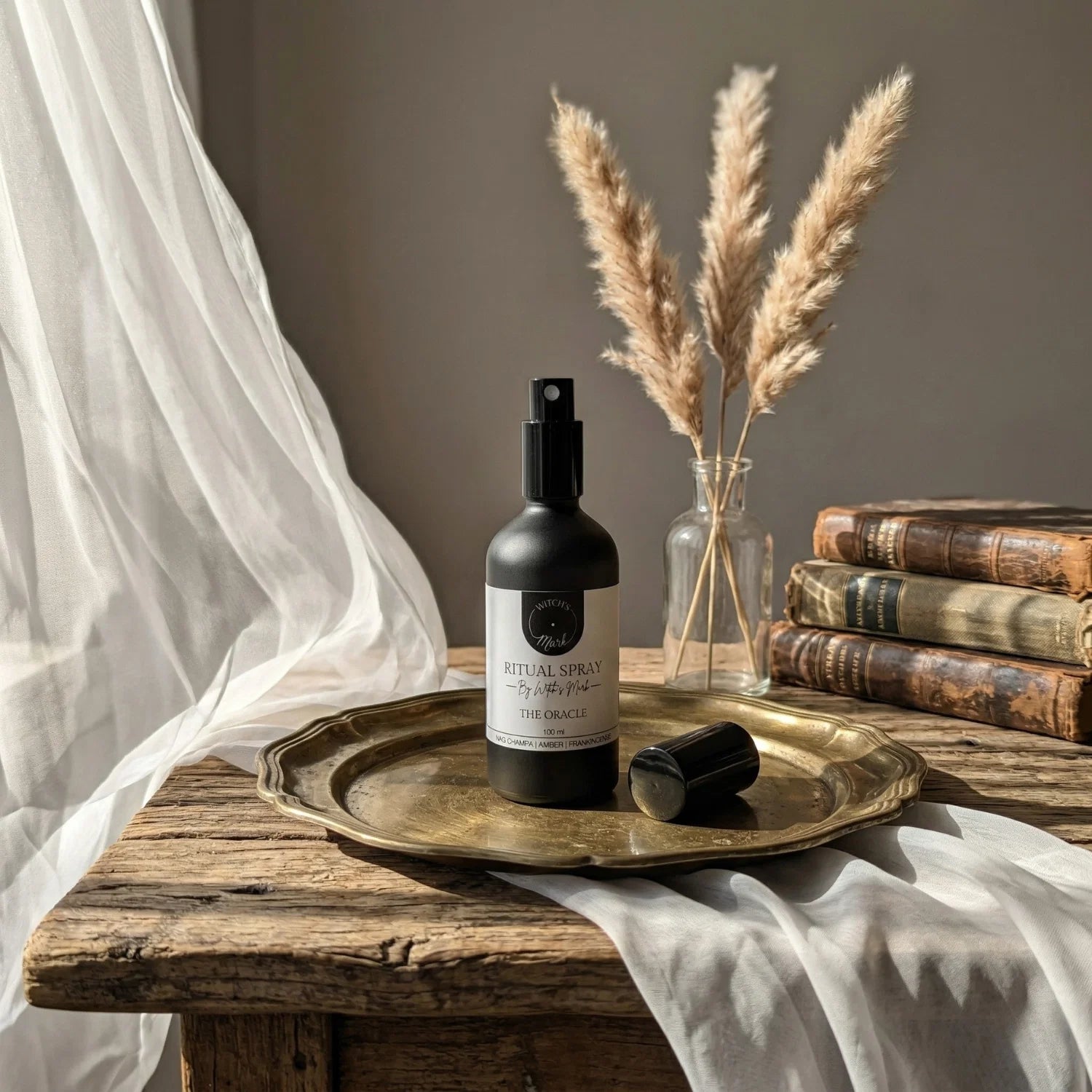 The matte black bottle of The Oracle ritual spray with its white label elegantly displayed on a vintage brass tray atop a rustic wooden table. This sacred elixir is perfect for misting over your altar or tarot deck to invoke absolute clarity and timeless insight.