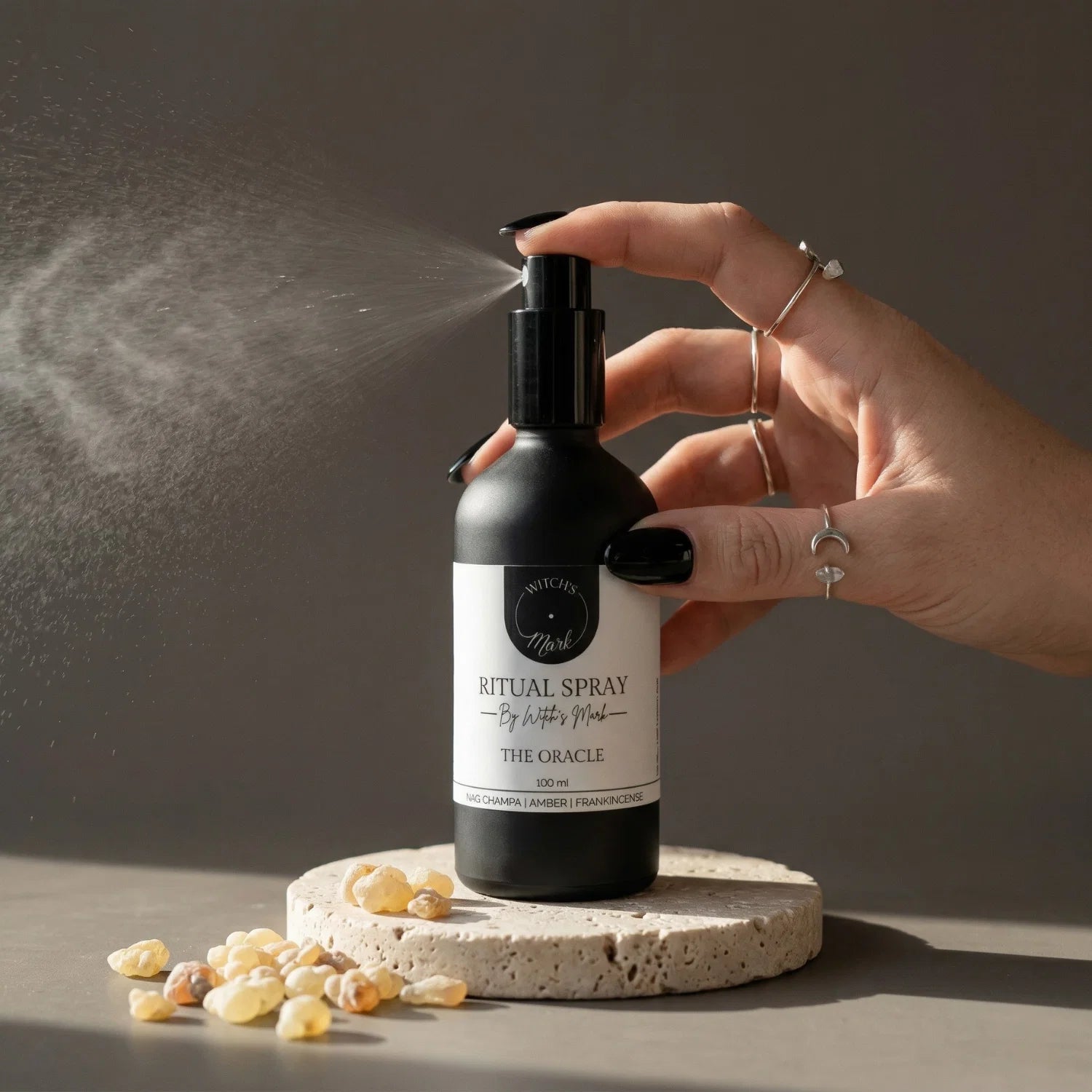 A hand with black nail polish spritzing the Witch's Mark The Oracle ritual spray over scattered frankincense tears. This illuminating aura mist is infused with citrine-charged moon water and rich resins to awaken inner wisdom and empower tarot divination.