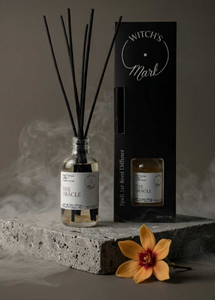 he Oracle spell jar reed diffuser by Witch's Mark displayed on a grey stone block next to its black packaging, surrounded by atmospheric swirling smoke and an orchid blossom.