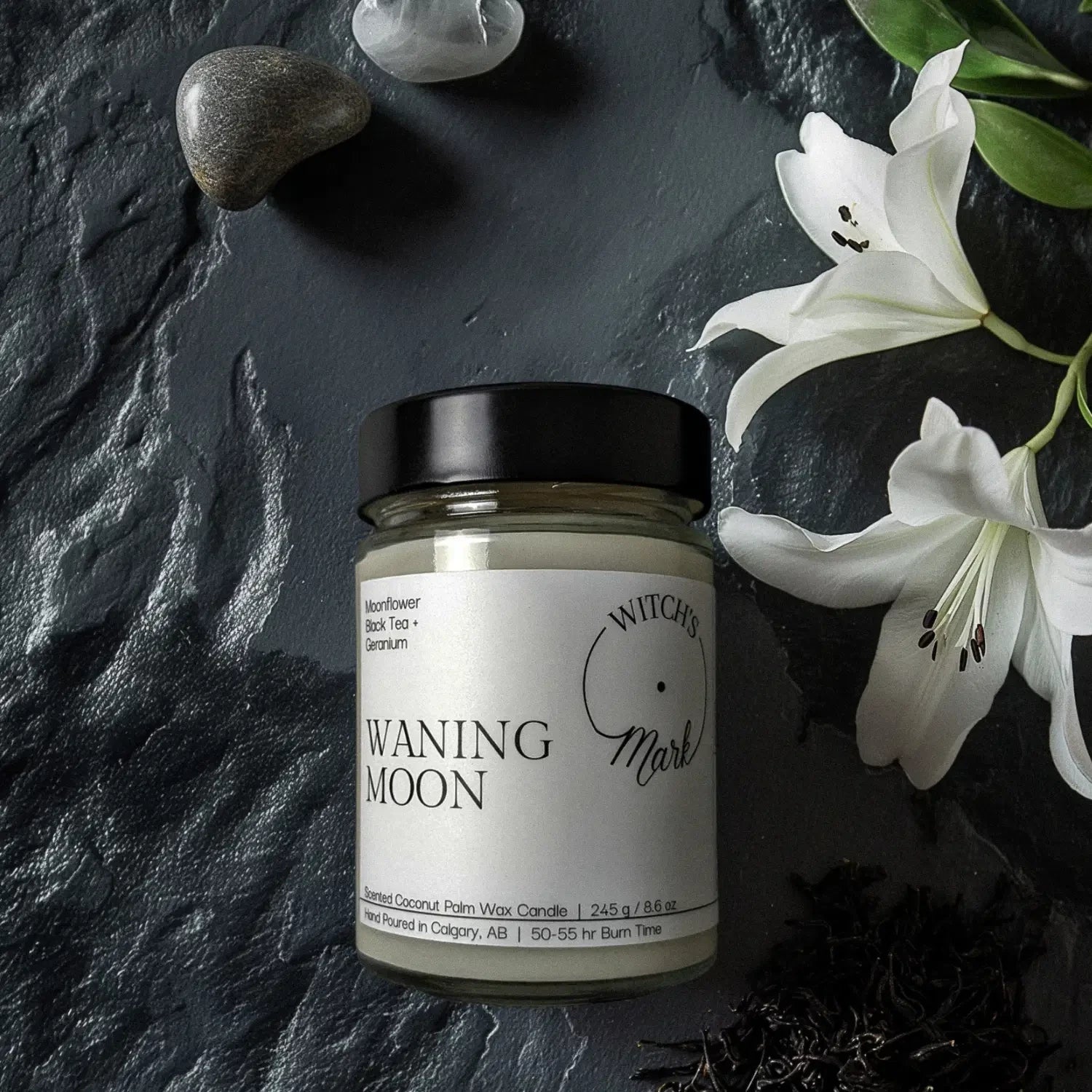 A top-down flat lay highlighting the ingredients of the Waning Moon candle, featuring the jar surrounded by blooming white moonflowers, loose black tea leaves, and smooth stones on a dark slate background.