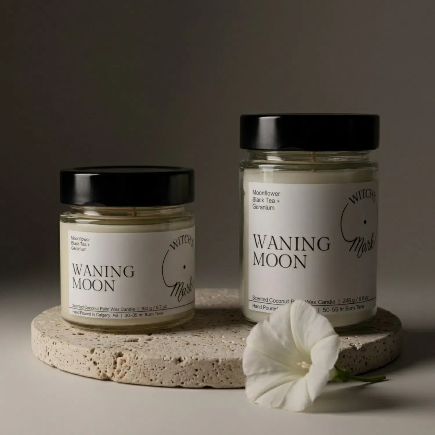 Two sizes of the Witch's Mark Waning Moon coconut wax candle, 5.7 oz and 8.6 oz, displayed on a round stone base and garnished with a delicate white moonflower.