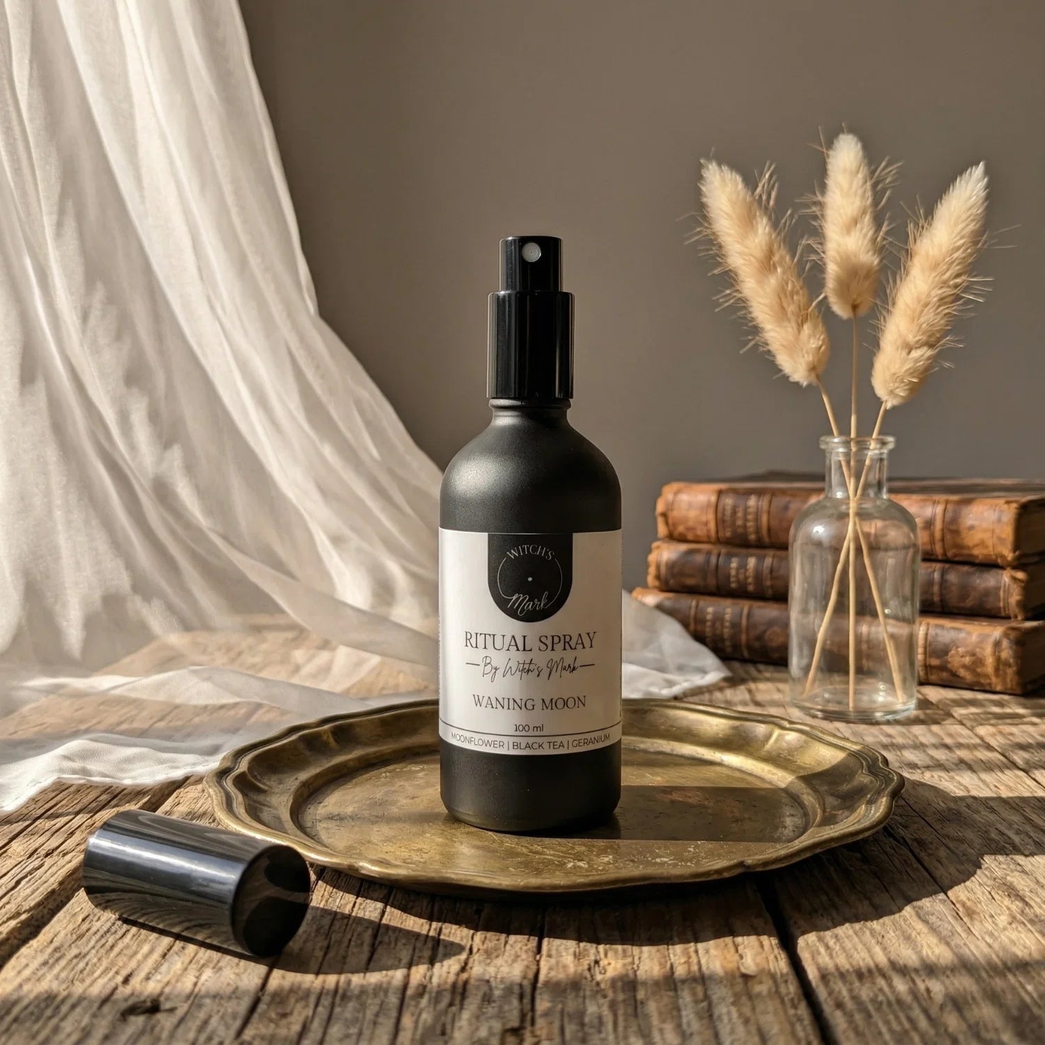 The matte black bottle of Waning Moon ritual spray with its white label elegantly displayed on a vintage brass tray atop a rustic wooden table bathed in soft light. This sacred elixir is perfect for misting over your altar to empower shadow work and banish stagnant energy.