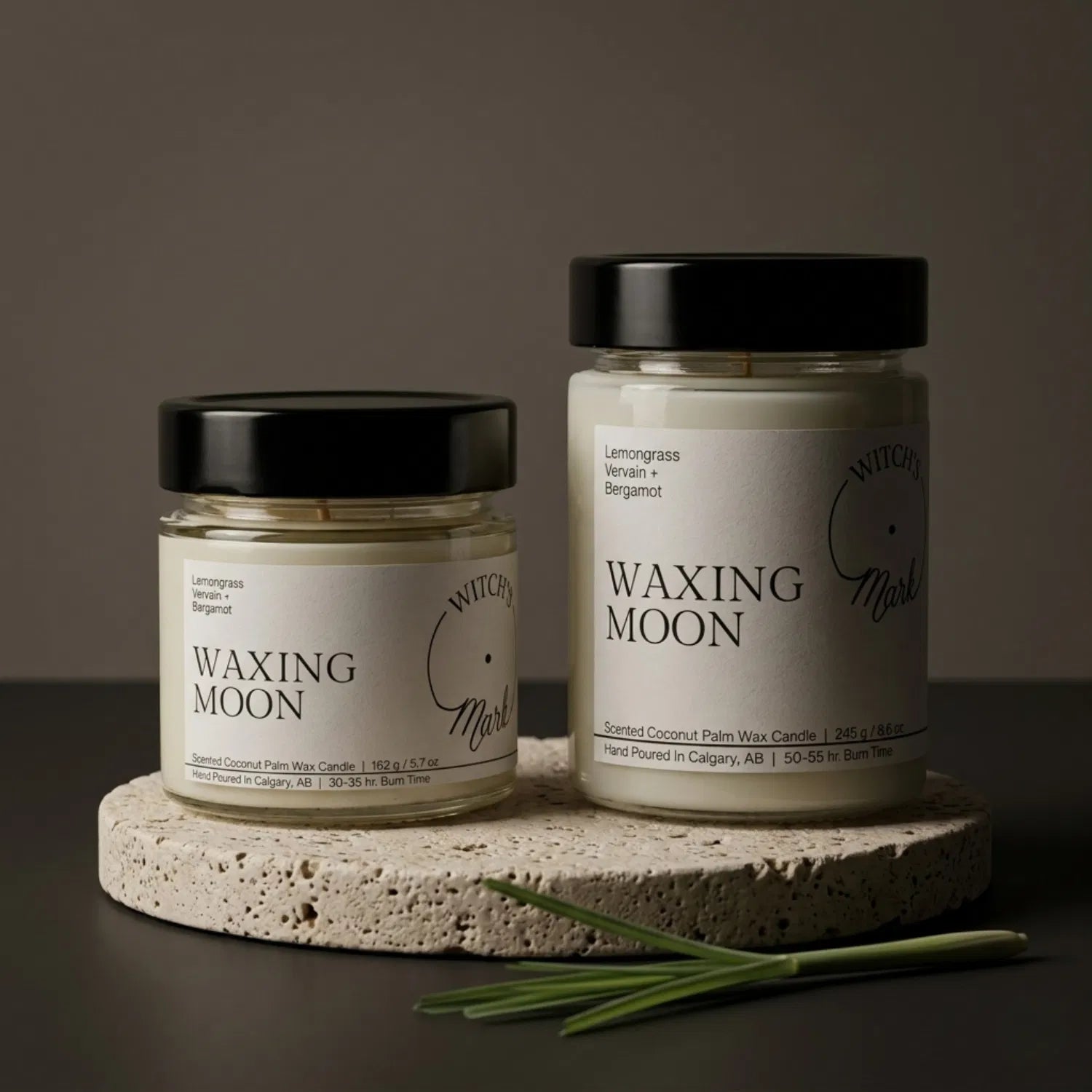 Two sizes of the Witch's Mark Waxing Moon coconut wax candle, 5.7 oz and 8.6 oz, displayed on a round stone base and garnished with fresh stalks of green lemongrass.