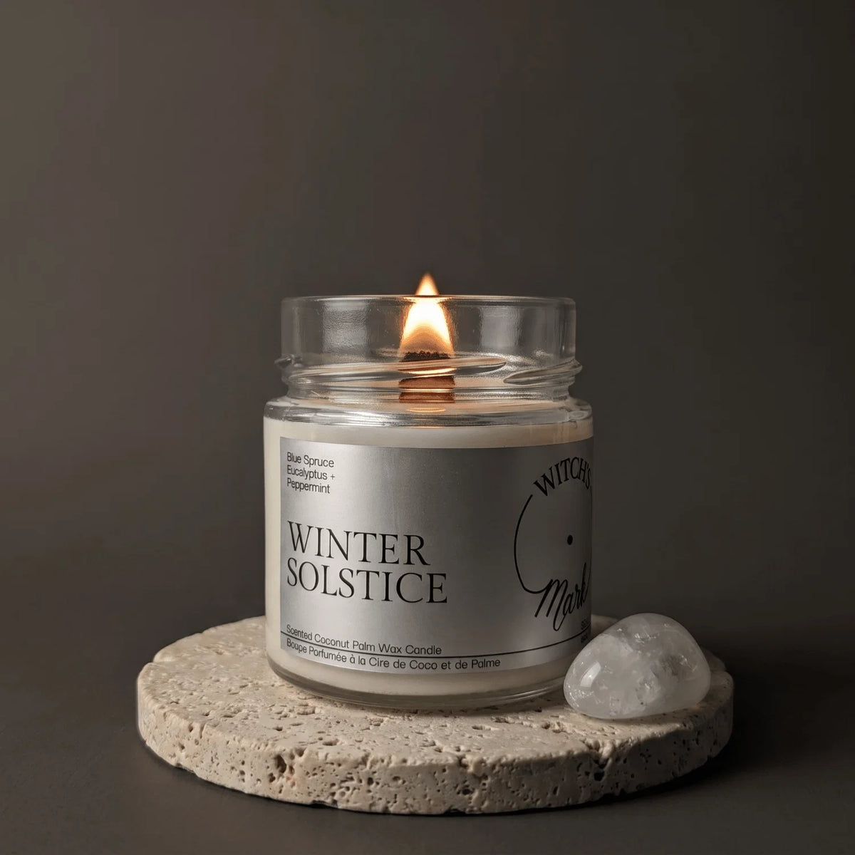 A lit candle in a clear glass jar with a white label reading 'WINTER SOLSTICE', placed on a round stone coaster with a snow quartz crystal beside it against a dark gray background.