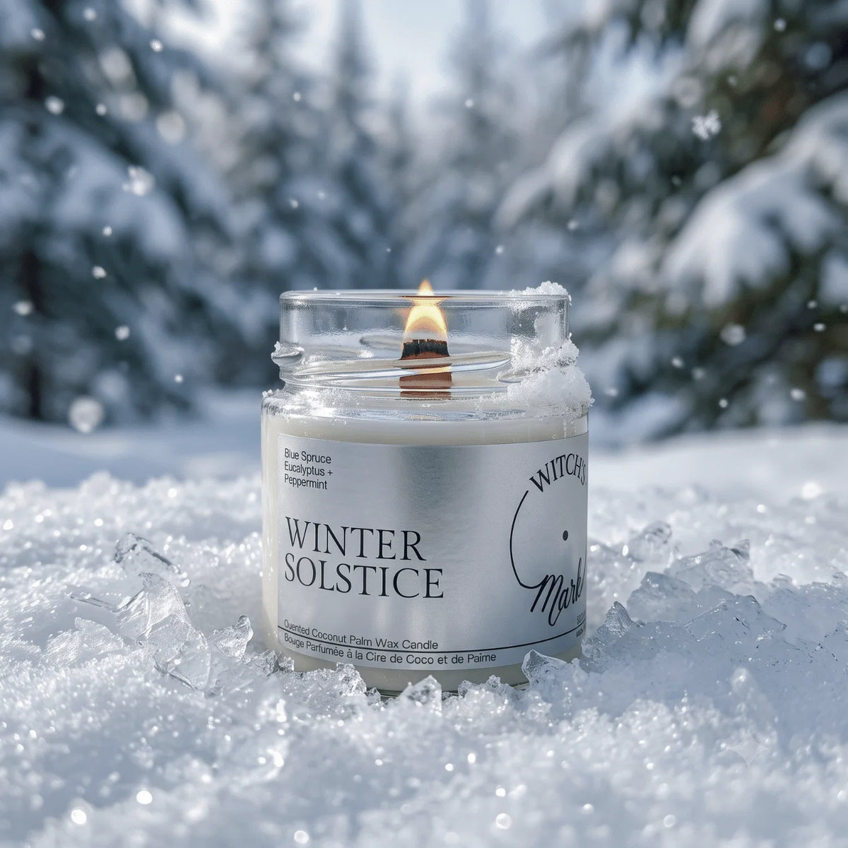A lit jar candle labeled "WINTER SOLSTICE" sits in snow outdoors, with blurred snowy trees in the background and falling snow visible around it.