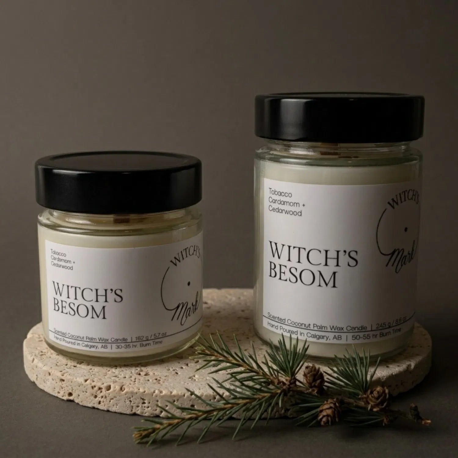 Two sizes of the Witch's Mark Witch's Besom coconut wax candle, 5.7 oz and 8.6 oz, displayed on a round stone base and garnished with a protective sprig of cedarwood.