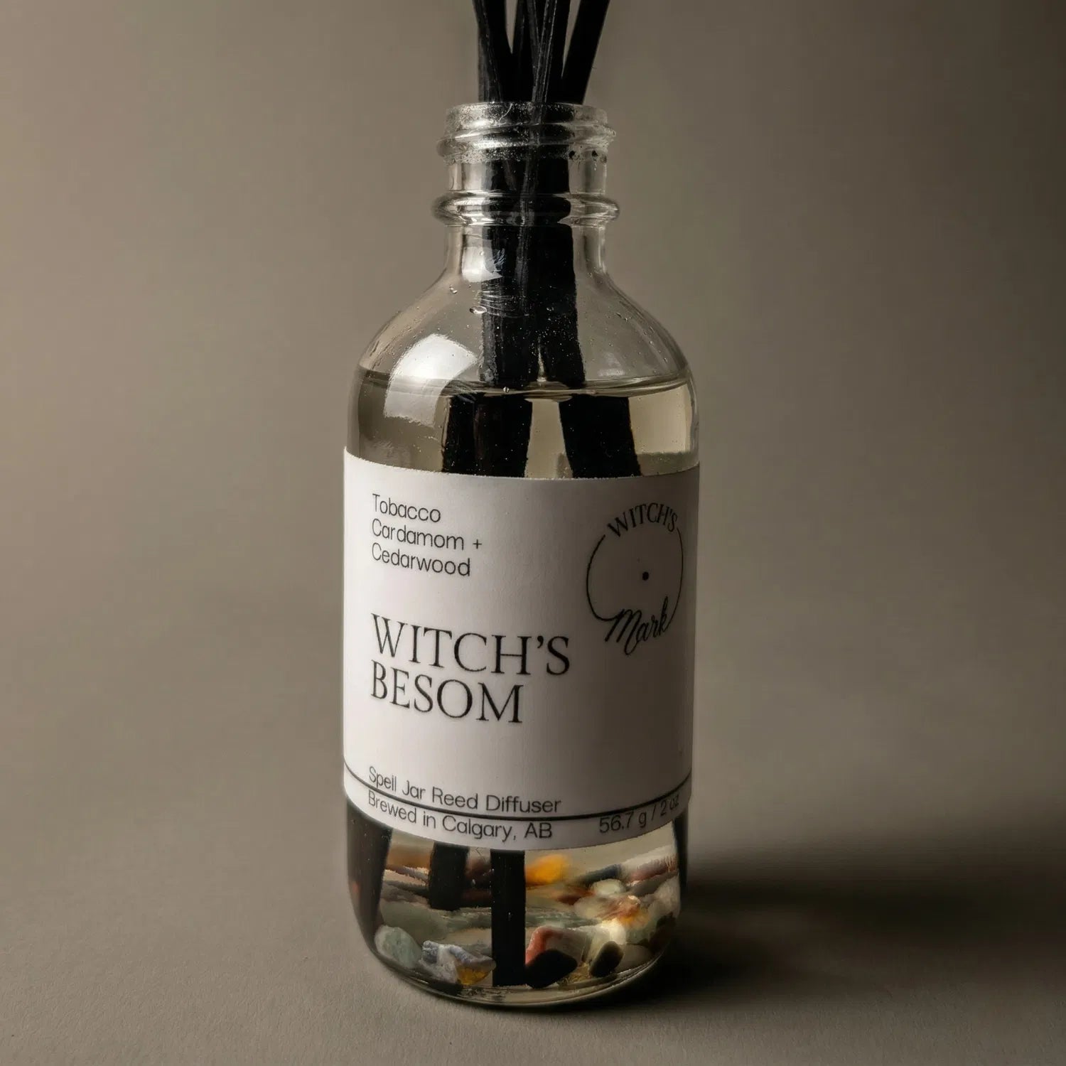 A dramatic spotlight close-up of the Witch's Besom spell jar reed diffuser, highlighting the front label which lists its magical, protective scent profile of grounding tobacco, cardamom, and deep resinous cedarwood.