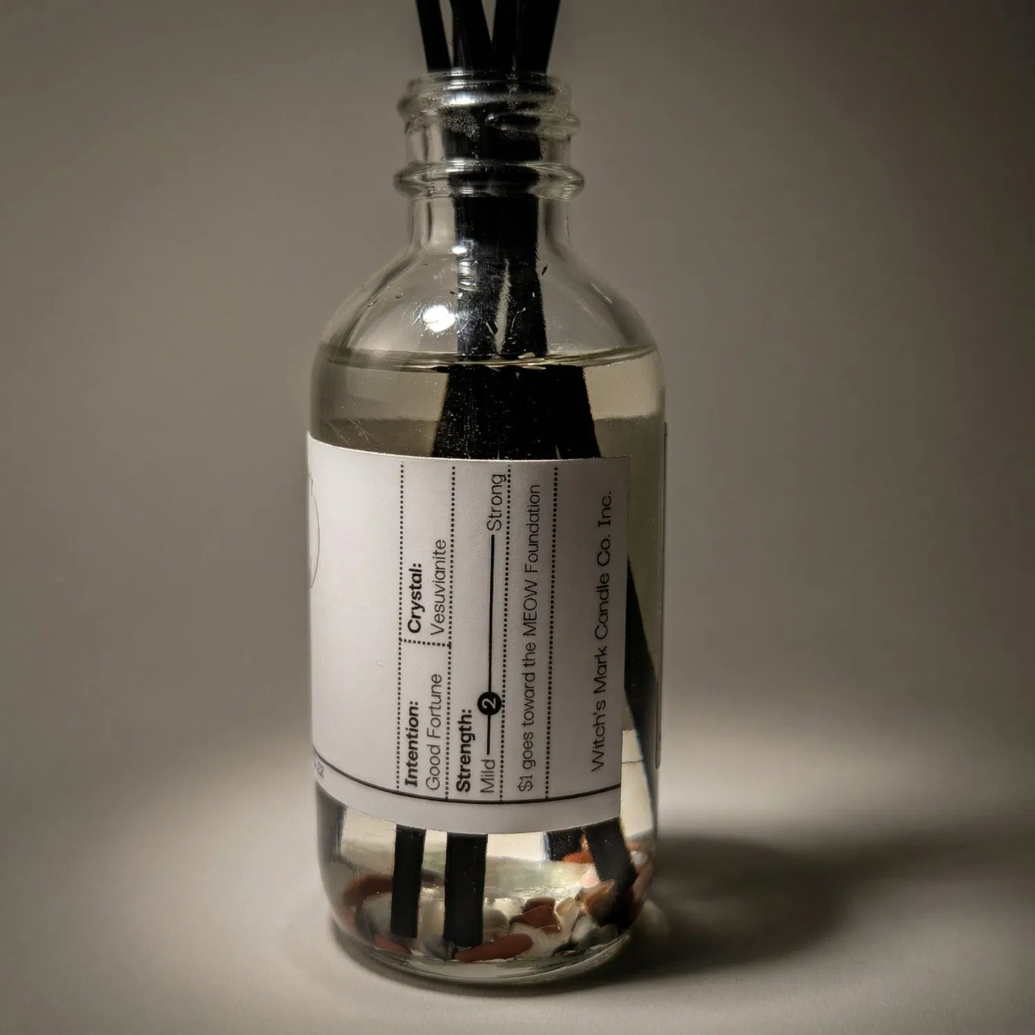 A warmly lit close-up of the Witch's Besom spell jar reed diffuser, revealing potent mixed crystals, including vesuvianite, submerged at the bottom of the clear botanical oil to help anchor turbulent emotions and balance the heart chakra.