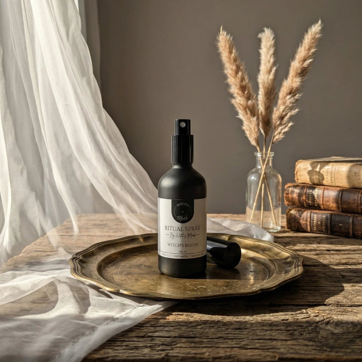 The matte black bottle of Witch's Besom ritual spray with its white label elegantly displayed on a vintage brass tray atop a rustic wooden table bathed in soft light. This sacred elixir is perfect for misting over your altar or threshold to welcome abundant blessings and draw in fierce prosperity.