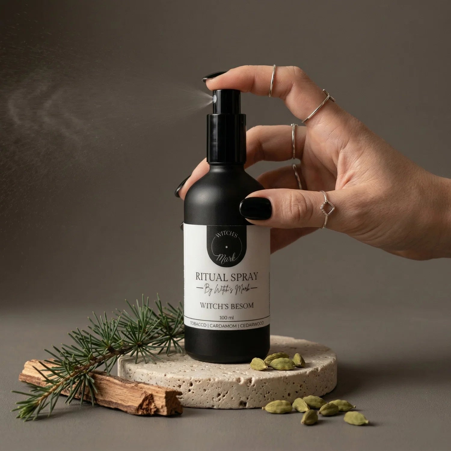 A hand with black nail polish spritzing the Witch's Mark Witch's Besom ritual spray over a fresh cedar branch, pieces of raw cedarwood, and green cardamom pods. This grounding aura mist is infused with jade-charged moon water to act as a sacred broom, sweeping away stagnant energy and energetic clutter.
