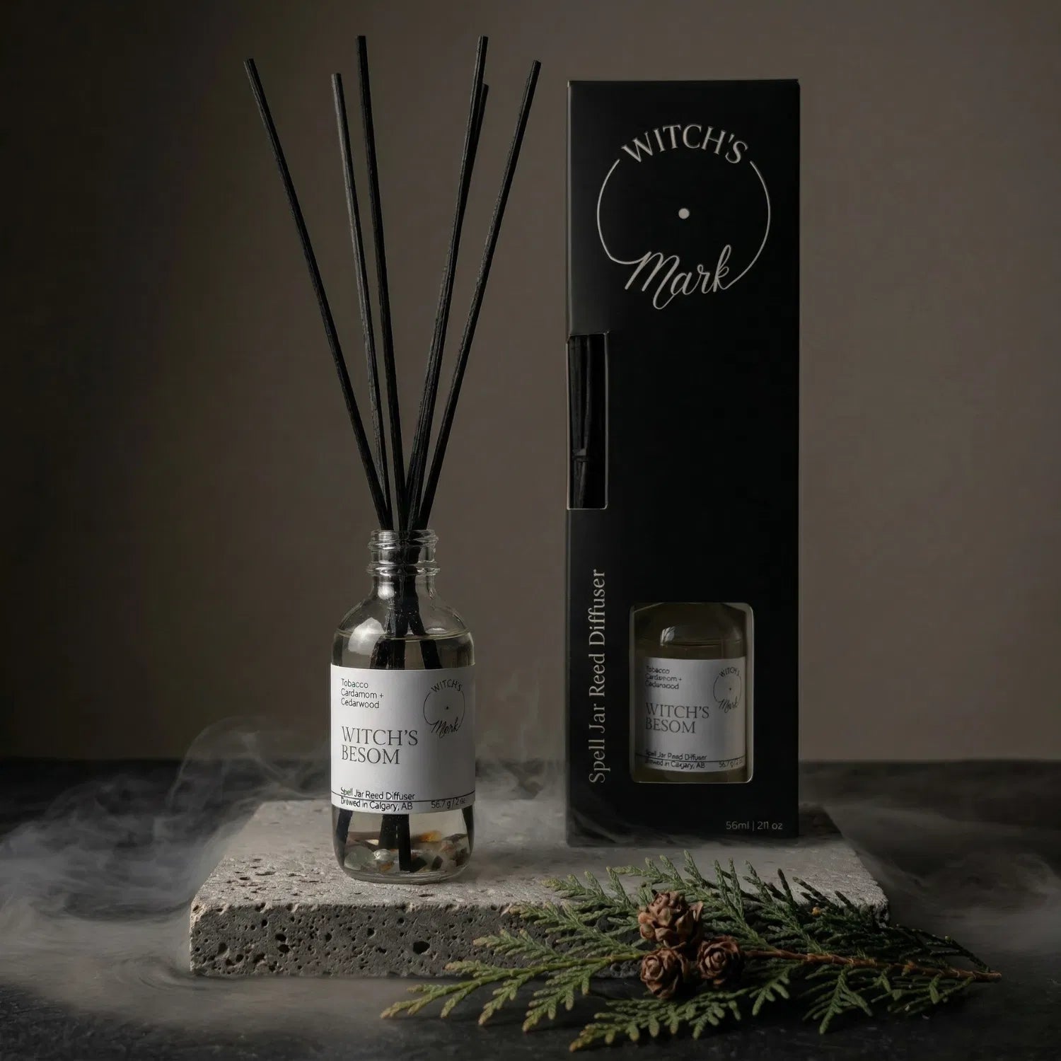 The Witch's Besom spell jar reed diffuser by Witch's Mark displayed next to its sleek black packaging box. The apothecary glass vessel with black reeds rests on a textured stone block surrounded by mystical mist and a fresh cedarwood sprig, serving as a perfect smokeless broom for space clearing.