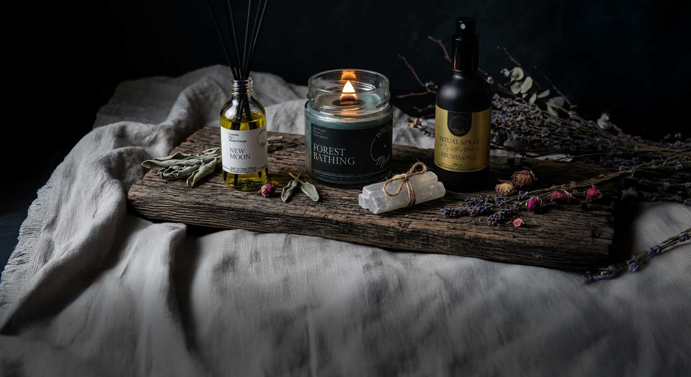 A moody display of Witch's Mark metaphysical products, featuring the New Moon reed diffuser, a lit Forest Bathing coconut wax candle, and an Abundance ritual spray on a rustic wooden board with a selenite crystal and dried botanicals.