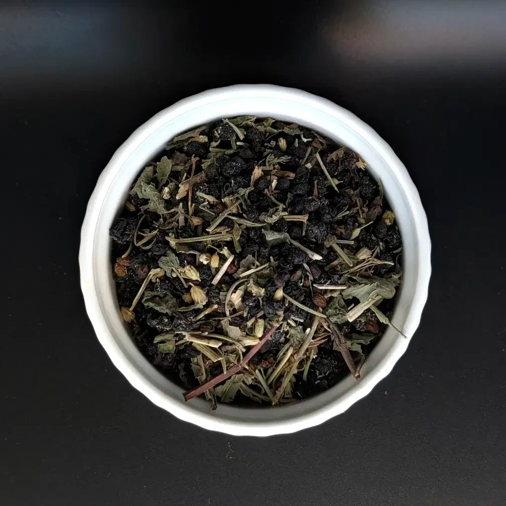 A top-down, close-up view of the Witch's Ward loose leaf herbal tea blend by Witch's Mark in a small white bowl. The image clearly showcases the high-quality mix of dark dried elderberries, cut rosemary, and restorative green herbal leaves against a dark background.